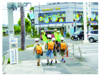 スクールガードの人たちが立つ歩道を黄色いランドセルを背負い通学帽子をかぶった児童たちが歩いている写真