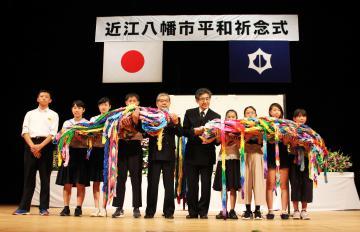 第9回近江八幡市平和記念式典にて、8,700羽の折鶴を掲げる小学生らと市長はが並んだ写真