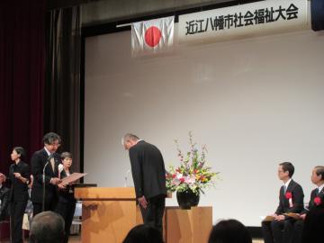 第9回近江八幡市社会福祉大会の開会式にて、表彰状を読み上げる市長と、お辞儀をする男性の写真