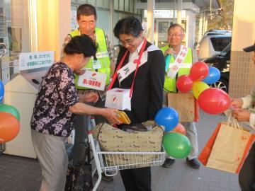 赤い羽根共同募金活動にて、募金箱を首から下げる市長と、募金を行う女性の写真