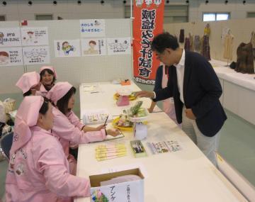 安土町地域自治区文化祭での健康推進課のブースにて、ピンク色の割烹着を着たスタッフから減塩味噌汁を受け取る市長の写真