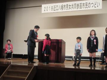 2018近江八幡市男女共同参画市民のつどいにて、壇上に立つ3名の小学生と、賞状を市長から授与される小学生の女の子の写真