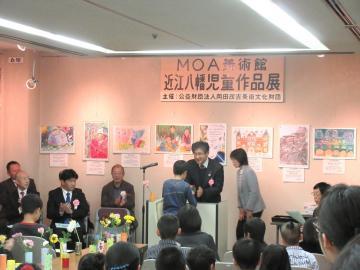 MOA美術館近江八幡児童作品展表彰式にて、表彰状を授与する市長と、受け取る児童、それらを着席し見守る人たちの写真