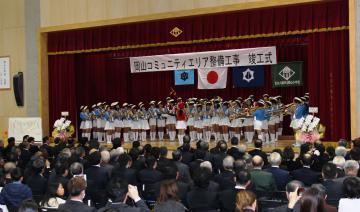 岡山小学校、岡山地域防災センターの竣工式にて、壇上での岡山小学校ブラスバンドの演奏を着席して聴く参加者らの写真