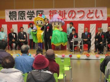 桐原学区福祉のつどいにて、壇上に並べられた椅子に着席するスーツの男性らと、その中央に立つ市長が着席する参加者に向かって話している写真