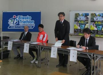 第5回びわ湖トライアスロンin近江八幡 記者会見にて、椅子に座るスーツの男性2名とユニフォームの男性の間に立って話す市長の写真