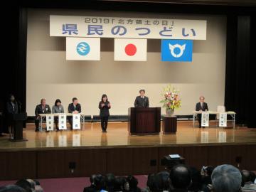 2019「北方領土の日」県民のつどいにて、壇上の端に立つ司会の女性と、椅子に座る4名の男女と、壇上の中央で話す市長の写真