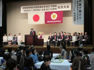 滋賀県地域女性団体連合会創立70周年・滋賀県婦人会館設立60周年記念大会にて着席した参加者と、壇上の椅子に座る参加者を前にマイクで話す市長の写真