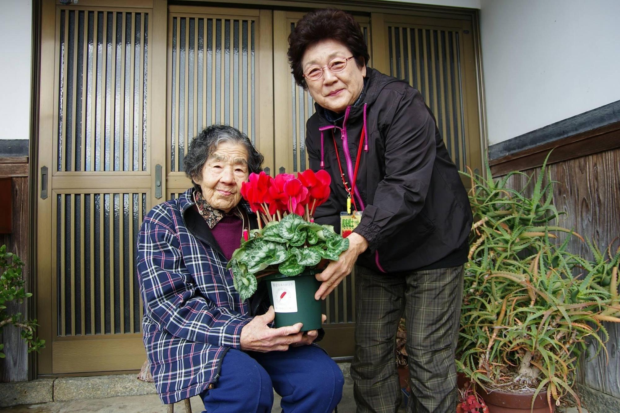 見守りの輪：玄関前にてシクラメンの植木鉢を持っている左に座っている女性、右に立っている女性の写真