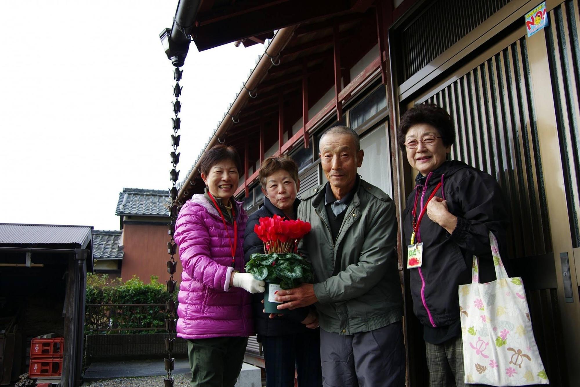 見守りの輪：玄関前にてシクラメンの植木鉢を持っている左から女性、女性、男性、女性が立っている写真