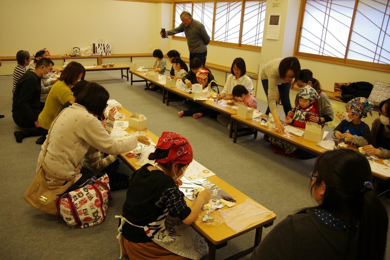 手をつなぐフェスタ：室内にてコの字テーブルで頭巾をかぶった子供たちと親たちが何かを作っている写真