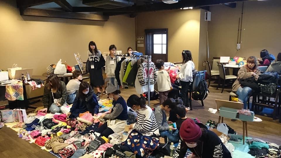 キッズフリマ：室内にて床に広げられた衣服などを座って整理している大人たち、後ろには立っている子供たちや衣装ハンガーラックが写っている写真