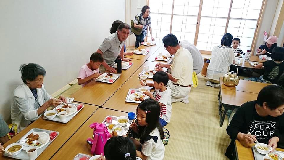 むさっ子食堂：長机の上に置かれた食事を食べている大人と子供たちの写真