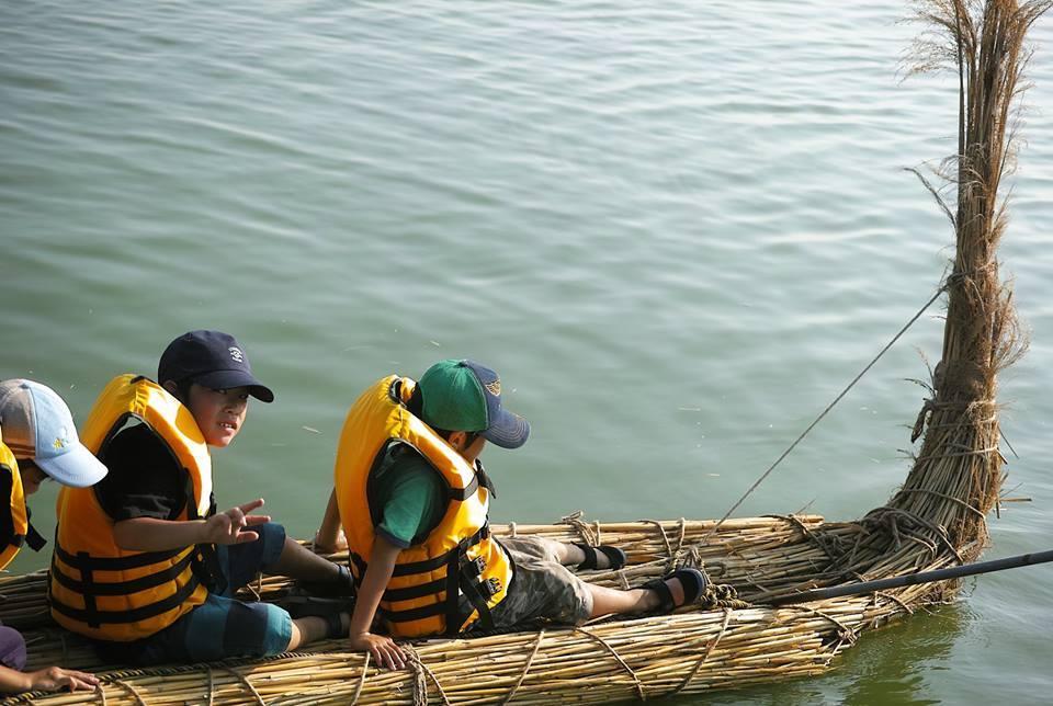 ヨシ船づくり：ヨシ船に乗って水の上に浮いている救命胴衣を着た子供の写真