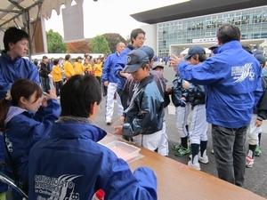 青いスタッフジャンパーを着たボランティアの皆さんが受付机や会場で少年野球チームの児童らを誘導している写真