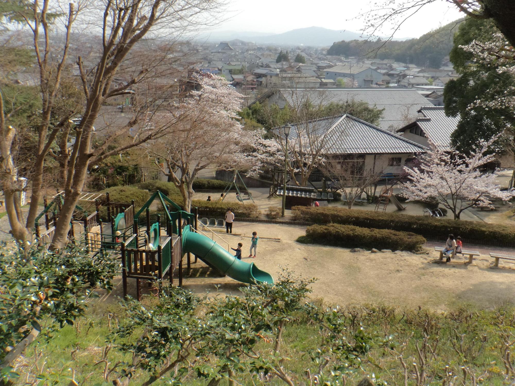 緑の滑り台のついた遊具で遊ぶ子供達とベンチに座っている大人達、桜の木が写っている八幡公園の写真