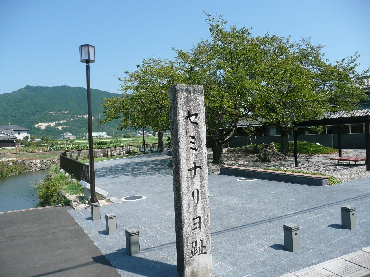 石の看板が立ち、背景にはなだらかな山の緑が青空に映えるセミナリヨ公園の木々やベンチの写真